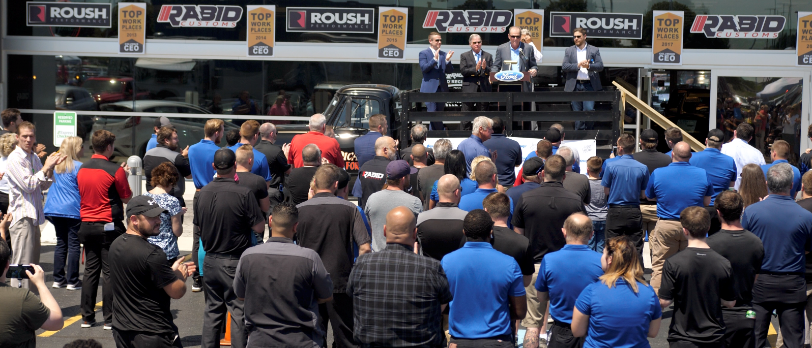 A diverse group of people stands together in front of a large building, smiling and engaging with one another.