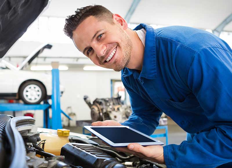 A mechanic examines his car while holding a tablet, focused on the device in his hands.